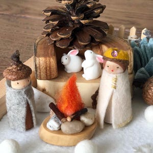 May include: A miniature woodland scene with a felt doll wearing a crown, two white bunnies, a small fire pit, and a pine cone.
