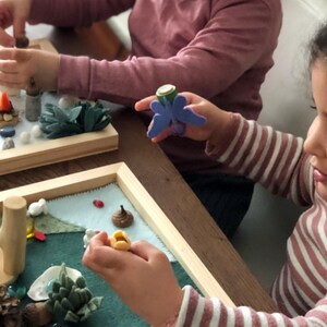 May include: Two children are playing with a felt nature scene in a wooden frame. The scene includes a variety of natural elements, such as leaves, rocks, and a small wooden tree. The children are using their hands to place the elements in the scene.