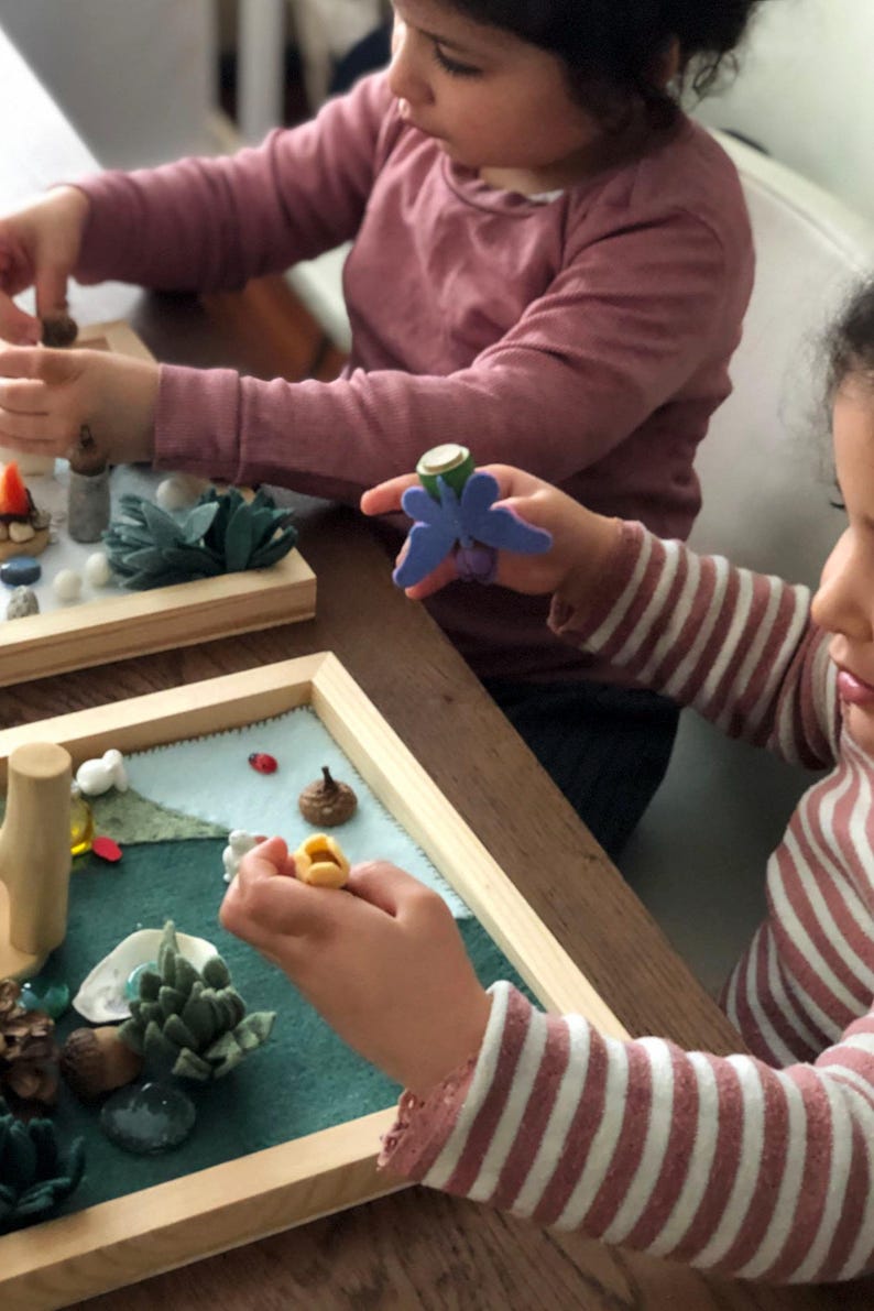 May include: Two children are playing with a felt nature scene in a wooden frame. The scene includes a variety of natural elements, such as leaves, rocks, and a small wooden tree. The children are using their hands to place the elements in the scene.