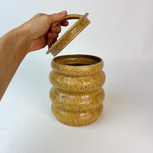 May include: A speckled yellow ceramic jar with a matching lid. The jar has a rounded, stacked design. The lid is being lifted, revealing the interior. The jar is a decorative storage container.