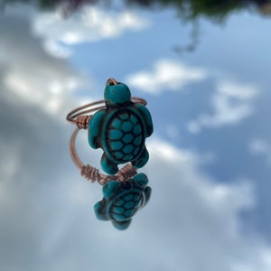 May include: A copper wire ring with a turquoise turtle charm. The ring is on a reflective surface.
