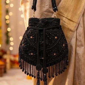 Peut inclure: Sac à main noir orné de perles avec des motifs floraux complexes et des franges pendantes. Le sac est doté d'une fermeture à cordon et d'éléments métalliques dorés. Il est tenu par une personne portant des bracelets dorés et un vêtement beige.