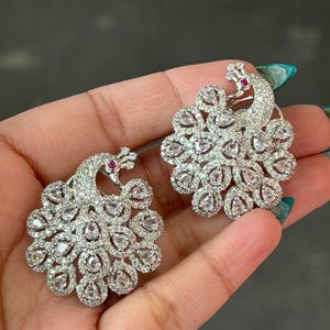 May include: A pair of silver-toned peacock earrings. Each earring features a peacock design with a fan of clear, teardrop-shaped stones and a small red stone at the head. The earrings are designed to be worn as studs.