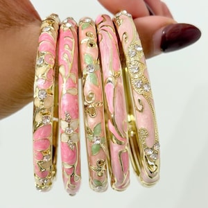 Op de afbeelding: Een verzameling van zes goudkleurige armbanden. Elke armband heeft een roze emaille bloemenontwerp met groene en witte accenten. Sommige armbanden zijn versierd met heldere kristallen. De armbanden worden in een hand gehouden.