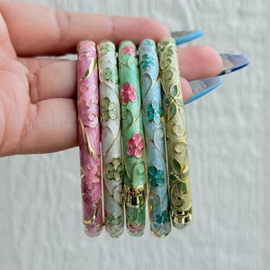 May include: Six enamel bangles in a variety of colours. Each bangle is decorated with a floral pattern in pink, green, blue, and gold. The bangles are held in a hand, highlighting their detailed design and bright colours.