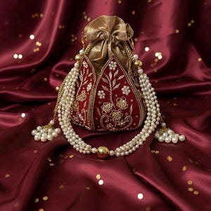 Może przedstawiać: Bordowa aksamitna torebka ze złotymi akcentami i kwiatowym haftem. Torebka ma zapięcie na sznurek i jest ozdobiona wielożyłowym naszyjnikiem z pereł ze złotymi koralikami i frędzlami. Tło to bordowa satynowa tkanina.