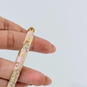 K&ouml;nnte beinhalten: Ein goldfarbener Armreif mit einer hellrosa Emaille-Einlage, verziert mit geschwungenen Mustern und eingebetteten klaren Kristallen. Der Metallrahmen hat ein poliertes Finish und das Design umfasst dekorative Ausschnitte entlang des Bandes.