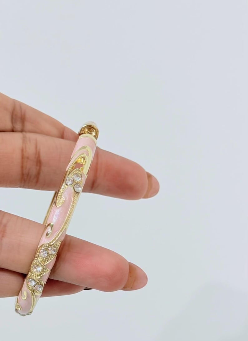 K&ouml;nnte beinhalten: Ein goldfarbener Armreif mit einer hellrosa Emaille-Einlage, verziert mit geschwungenen Mustern und eingebetteten klaren Kristallen. Der Metallrahmen hat ein poliertes Finish und das Design umfasst dekorative Ausschnitte entlang des Bandes.
