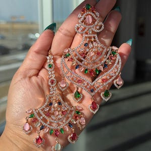 Könnte beinhalten: Ein Paar roségoldfarbene Chandelier-Ohrringe mit klaren Kristallen und farbigen Edelsteinen. Die Ohrringe haben rosa, grüne, blaue und rote Steine, ein mehrschichtiges Design und tropfenförmige Anhänger. Ein elegantes Accessoire.