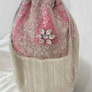 May include: Pink drawstring bag with silver beaded detailing and a flower-shaped jewel. The bag has a long fringe of white beads and a silver drawstring. A fashionable accessory.