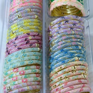 May include: Stacks of colorful enamel bangles in clear plastic containers. The bangles feature floral and abstract designs in shades of pink, blue, green, yellow, and purple, with gold-colored metal accents. The bracelets are stacked neatly, showcasing a variety of patterns.