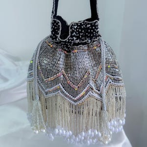May include: A silver and white beaded drawstring bag with a black velvet top. The bag has a zigzag pattern with iridescent sequins and a fringe of white beads with pearl accents. The bag is a stylish accessory.