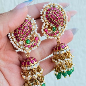 May include: Gold earrings with a paisley design, featuring red and green gemstones, and white pearls. The earrings have a dangling bell shape with green beads and white pearls.