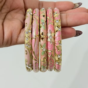 K&ouml;nnte beinhalten: Ein Set aus f&uuml;nf goldfarbenen Armreifen mit floralen Emaille-Mustern in Rosa und Hellgr&uuml;n. Jeder Armreif ist mit kleinen, klaren, runden Strasssteinen verziert, die in die filigranen Metallverzierungen eingelassen sind.