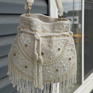 Peut inclure: Sac à main ivoire perlé avec fermeture à cordon et bandoulière. Le sac est orné de perles, de perles transparentes et d'accents dorés. Le bas du sac est orné d'une frange de perles transparentes.
