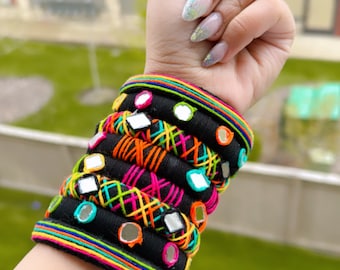 May include: A stack of black fabric bracelets adorned with colourful embroidery and small mirrored details. The bracelets feature vibrant stitching in orange, pink, and green, creating a contrast against the black. The bracelets are stacked on a wrist.