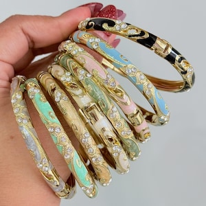 May include: A selection of gold-coloured bangles with intricate patterns. Each bangle displays a different enamel colour, such as black, blue, pink, and green, embellished with clear rhinestones. The bangles are held together, highlighting their decorative features.