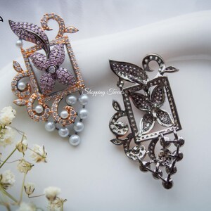 Puede incluir: Dos broches ornamentados con diseños florales y de hojas. Uno es de oro rosa con piedras rosas y transparentes, el otro es plateado con piedras negras. Ambos tienen detalles de perlas y el texto "Shopping Tweets".
