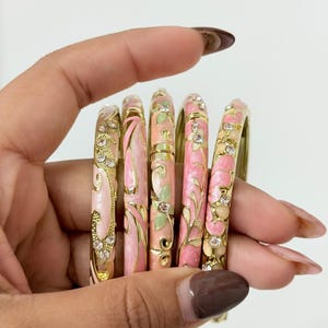 K&ouml;nnte beinhalten: Ein Set aus f&uuml;nf goldfarbenen Armreifen mit floralen Mustern aus rosa und hellgr&uuml;nem Emaille sowie eingelegten klaren Kristallen. Jeder Armreif verf&uuml;gt &uuml;ber ein Klappdesign mit filigranen, geschwungenen Details.