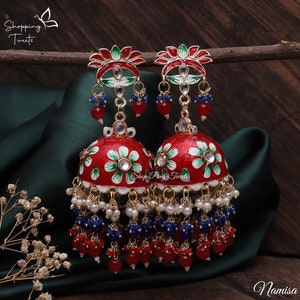 May include: A pair of red and white floral earrings with dangling red and blue beads. The earrings have a bell shape with intricate detailing and are adorned with small white pearls. The earrings are displayed on a dark green fabric.