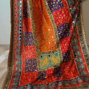 May include: A colourful patchwork textile in shades of red, orange, blue, and gold. The fabric is decorated with detailed patterns, mirrored embellishments, and a decorative border. The design combines traditional and ornate elements, creating a visually rich piece.
