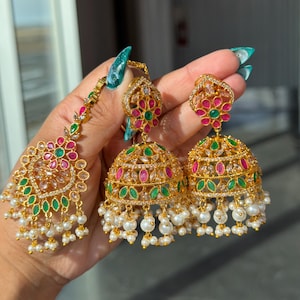 May include: Gold-toned jewelry set featuring a headpiece and earrings. The headpiece and earrings have a floral design with red, green, and clear gemstones. The earrings are bell-shaped and adorned with white pearls. The jewelry is held up by a hand.