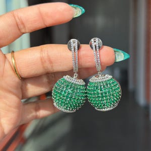 May include: A pair of dangling earrings featuring a round, emerald green design, accented with small, sparkling white stones. The earrings have a silver-colored post and a delicate, elongated chain.