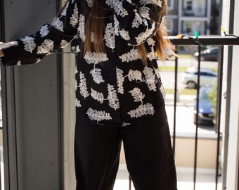 May include: A black shirt with white leaf embroidery and black wide-leg trousers. The outfit is worn on a balcony. The shirt has long sleeves and a classic collar. The outfit is suitable for casual wear.
