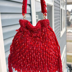 May include: A vibrant red beaded handbag with a drawstring closure. The bag features a textured, woven design and is adorned with dangling tassels. The straps are a matching red and attached with gold-colored hardware. A stylish accessory.