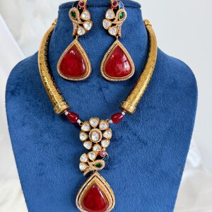 May include: A gold-toned necklace and earring set featuring large, teardrop-shaped red stones. The necklace has a textured, rigid collar with a central floral pendant adorned with clear stones, small red and green accents, and a dangling red teardrop.