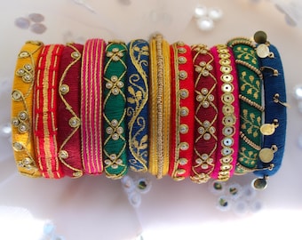 May include: A selection of vibrant bangles in shades of yellow, red, pink, green, and blue. The bangles are decorated with gold-coloured embellishments, embroidery, and small decorative details. The bangles are likely made of fabric and are designed for wear.