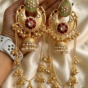 May include: Ornate gold-coloured earrings with detailed patterns. Each earring has a green, teardrop-shaped stone, a red flower design, and hanging chains with small gold beads. The earrings are designed in a traditional style.