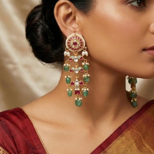 May include: Gold chandelier earrings with a circular top featuring red and white accents. The earrings have multiple dangling strands with green and red gemstones and white pearls. The earrings are worn by a person wearing a red and gold garment.