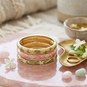 May include: Stacked gold bangles with floral enamel designs in pink, green, and white. The bangles are on a pink surface with decorative elements, including a small gold dish, ring, and gemstones. The overall theme is jewelry and accessories.
