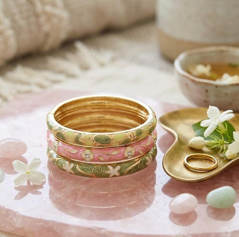 May include: Stacked gold bangles with floral enamel designs in pink, green, and white. The bangles are on a pink surface with decorative elements, including a small gold dish, ring, and gemstones. The overall theme is jewelry and accessories.