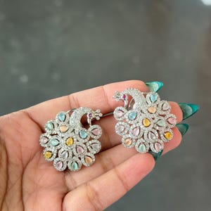 Op de afbeelding: Een paar zilverkleurige pauwoorbellen. Elke oorbel heeft een pauwontwerp met een lichaam van kleurrijke edelstenen in pasteltinten van roze, geel, blauw en groen. De kop van de pauw is versierd met een rode edelsteen.