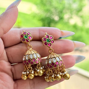 Puede incluir: Pendientes dorados con un diseño floral en la parte superior, con piedras preciosas rosas y verdes. Los colgantes en forma de campana están adornados con detalles blancos y rosas, pequeñas cuentas blancas y abalorios dorados. Los pendientes se sostienen en una mano.