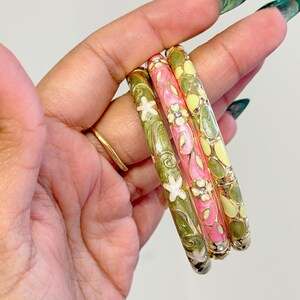 May include: Three colorful enamel bangles. One is pink with white flowers and clear gems, another is green with white flowers, and the third is yellow with green accents. The bangles are decorative and have a floral design.