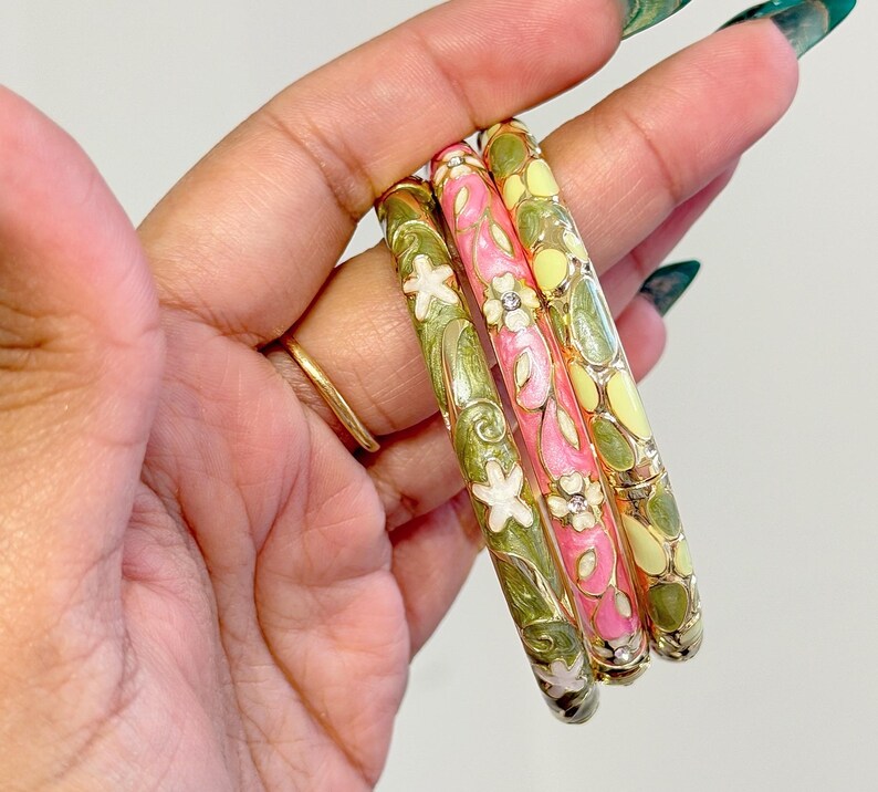 May include: Three colorful enamel bangles. One is pink with white flowers and clear gems, another is green with white flowers, and the third is yellow with green accents. The bangles are decorative and have a floral design.