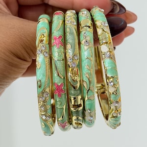 May include: A collection of five hinged bangles. Each bangle features a light green enamel base with gold-colored accents and floral designs. The bangles are adorned with clear rhinestones and pink flower accents.