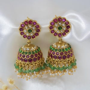 May include: A pair of gold earrings with a floral top and a bell-shaped base. The earrings are adorned with red, green, and white gemstones. Small green beads and white pearls hang from the bottom of the earrings.