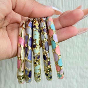 May include: A collection of six colorful enamel bangles with gold-tone metal accents. Each bangle features a unique pattern of pastel colors, including pink, purple, blue, and green, with small crystal embellishments. The bangles are designed with a mosaic-like pattern.