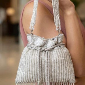 May include: A white beaded drawstring bag with a silver chain strap. The bag features vertical rows of beads, a beaded fringe, and decorative tassels. The bag is a formal accessory.