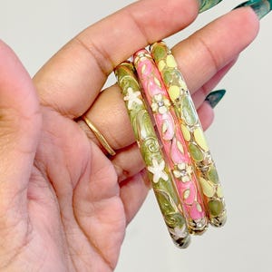 May include: Three colorful enamel bangles. One is pink with white flowers and clear rhinestones, another is green with white flowers, and the third is yellow with green accents. The bangles are held in a hand.