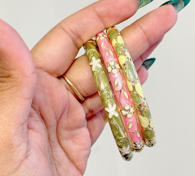 May include: Three colorful enamel bangles. One is pink with white flowers and clear rhinestones, another is green with white flowers, and the third is yellow with green accents. The bangles are held in a hand.