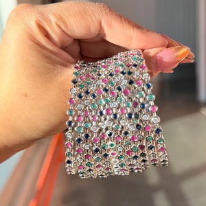May include: A collection of silver bracelets with colourful gemstones. Each bracelet features a series of round, multi-coloured gems, including pink, blue, green, and clear stones. The bracelets are held together by a silver chain.