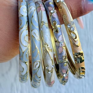 Könnte beinhalten: Set mit sechs dekorativen Armreifen in verschiedenen Farben, darunter Hellblau, Weiß und Gold. Die Armreifen weisen Blumen- und Wirbelmuster mit Strassakzenten auf. Der Text "Set 8" ist sichtbar.