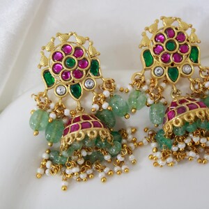 Peut inclure: Une paire de boucles d'oreilles dorées avec un motif floral, ornée de pierres précieuses roses et vertes. Les boucles d'oreilles ont des perles vertes pendantes et de petites perles blanches.