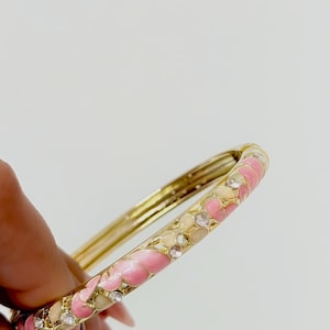 K&ouml;nnte beinhalten: Ein goldfarbener Klapparmreif mit einem Emaillemuster aus abwechselnden rosa und cremefarbenen Blumenformen, akzentuiert durch kleine, klare, runde Strasssteine, die in die Au&szlig;enfl&auml;che des Metallbandes eingelassen sind.