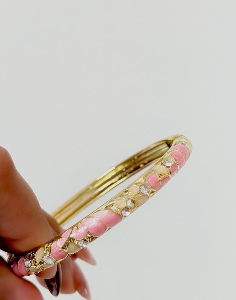 K&ouml;nnte beinhalten: Ein goldfarbener Klapparmreif mit einem Emaillemuster aus abwechselnden rosa und cremefarbenen Blumenformen, akzentuiert durch kleine, klare, runde Strasssteine, die in die Au&szlig;enfl&auml;che des Metallbandes eingelassen sind.